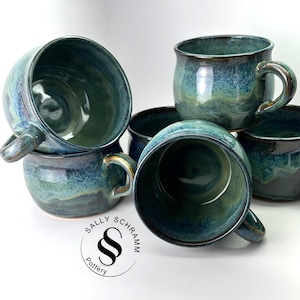 May include: A collection of handmade ceramic mugs in shades of teal and blue. Each mug features a rounded body, a sturdy handle, and a glossy finish. The mugs are stacked and arranged, showcasing their unique glaze patterns and textures. The logo "SALLY SCHRAMM Pottery" is visible.