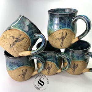 Puede incluir: Tazas de cerámica hechas a mano con un esmalte azul y verde. Cada taza presenta un diseño de colibrí y una base beige. Las tazas tienen asas grandes y están apiladas. Son de Sally Schramm Pottery.