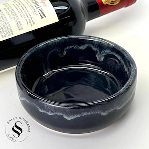 Puede incluir: Un cuenco de cerámica azul oscuro con un acabado brillante y un borde blanco. El cuenco es redondo y está sobre una superficie blanca. Una botella de vino tinto está en el fondo.