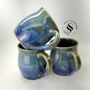 May include: Three ceramic mugs with a glossy glaze in shades of blue, green, and grey. The mugs feature a rounded shape and curved handles. A circular logo with the text Sally Schramm Pottery is visible in the upper right corner of the image.