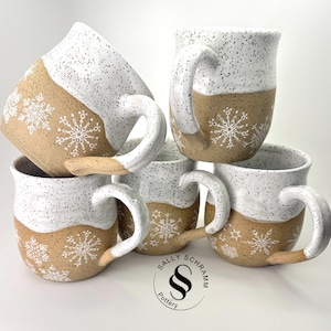 May include: A stack of six ceramic mugs. The mugs are white with black speckles on the top half and tan on the bottom half. White snowflake designs are painted on the tan sections. Each mug has a large, curved handle. The logo "Sally Schramm Pottery" is visible.