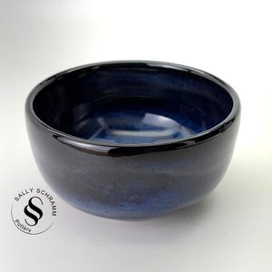 May include: A round ceramic bowl with a glossy finish, featuring a deep navy blue and black gradient glaze. The interior is a lighter shade of blue, transitioning to dark tones. The text SALLY SCHRAMM Pottery is printed in black on the lower left.