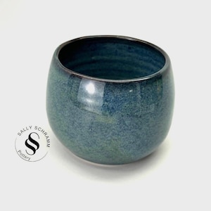 Puede incluir: Un cuenco de cerámica hecho a mano con forma redondeada y un esmalte azul-verdoso brillante y moteado. El borde es de un tono azul más oscuro. El cuenco está marcado con el texto "SALLY SCHRAMM Pottery".