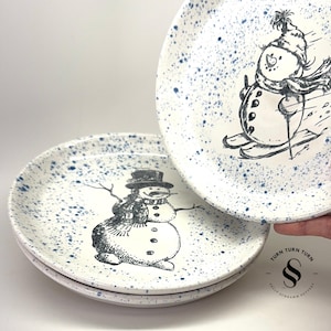 May include: Set of three white ceramic dessert plates with blue speckles. Each plate features a black and white illustration of a snowman. One snowman is skiing and the other is standing. The plates are stacked on top of each other. The bottom plate has the text "Turn Turn Turn Sally Schramm Pottery" printed on the bottom.