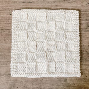 May include: A square, off-white knitted washcloth with a checkerboard pattern. The washcloth is made of a soft, textured yarn and has a simple, clean design. The washcloth is approximately 8 inches by 8 inches.