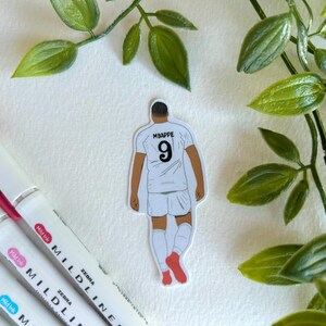 May include: A sticker featuring a soccer player in a white uniform with the name "MBAPPE" and the number "9" on the back. The sticker has a white background and is surrounded by green leaves and colorful markers.