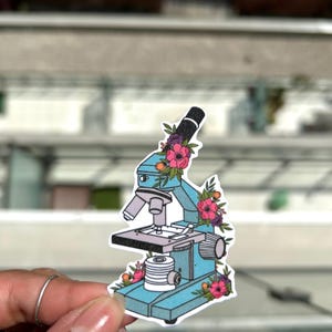 Peut inclure: Un microscope bleu avec un motif floral. Le microscope a un bras noir et une base argentée. Les fleurs sont roses, violettes et blanches.
