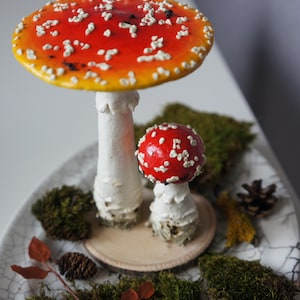 May include: Two decorative mushroom figurines with red and white caps and white stems. The larger mushroom has a wide cap with white spots. The mushrooms are on a decorative plate with moss and a small pine cone.