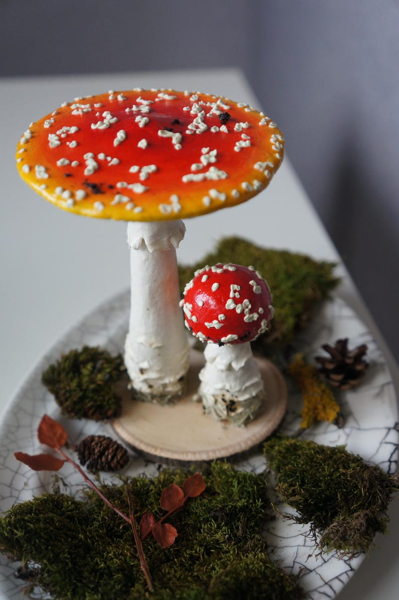 May include: Two decorative mushroom figurines with red and white caps and white stems. The larger mushroom has a wide cap with white spots. The mushrooms are on a decorative plate with moss and a small pine cone.