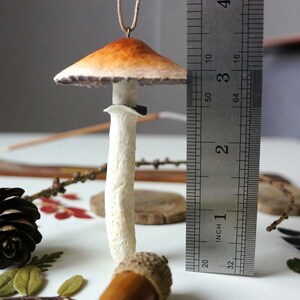 Może przedstawiać: Ręcznie robiony ceramiczny ornament w kształcie grzyba z brązowo-pomarańczowym kapeluszem i białym trzonem. Ornament ma około 5 cm wysokości i jest zawieszony na brązowym sznurku.