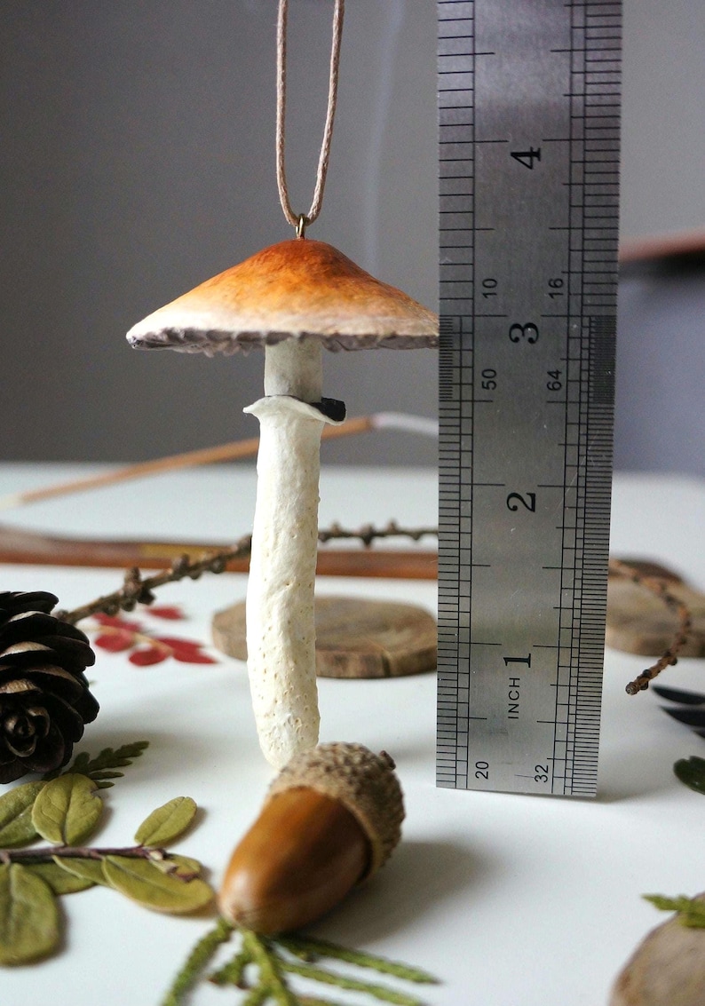 Może przedstawiać: Ręcznie robiony ceramiczny ornament w kształcie grzyba z brązowo-pomarańczowym kapeluszem i białym trzonem. Ornament ma około 5 cm wysokości i jest zawieszony na brązowym sznurku.