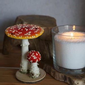 May include: Two decorative mushroom figurines with red caps and white spots, one large and one small, sit on a wooden surface. A lit candle in a glass jar is also present, creating a cozy, natural aesthetic.