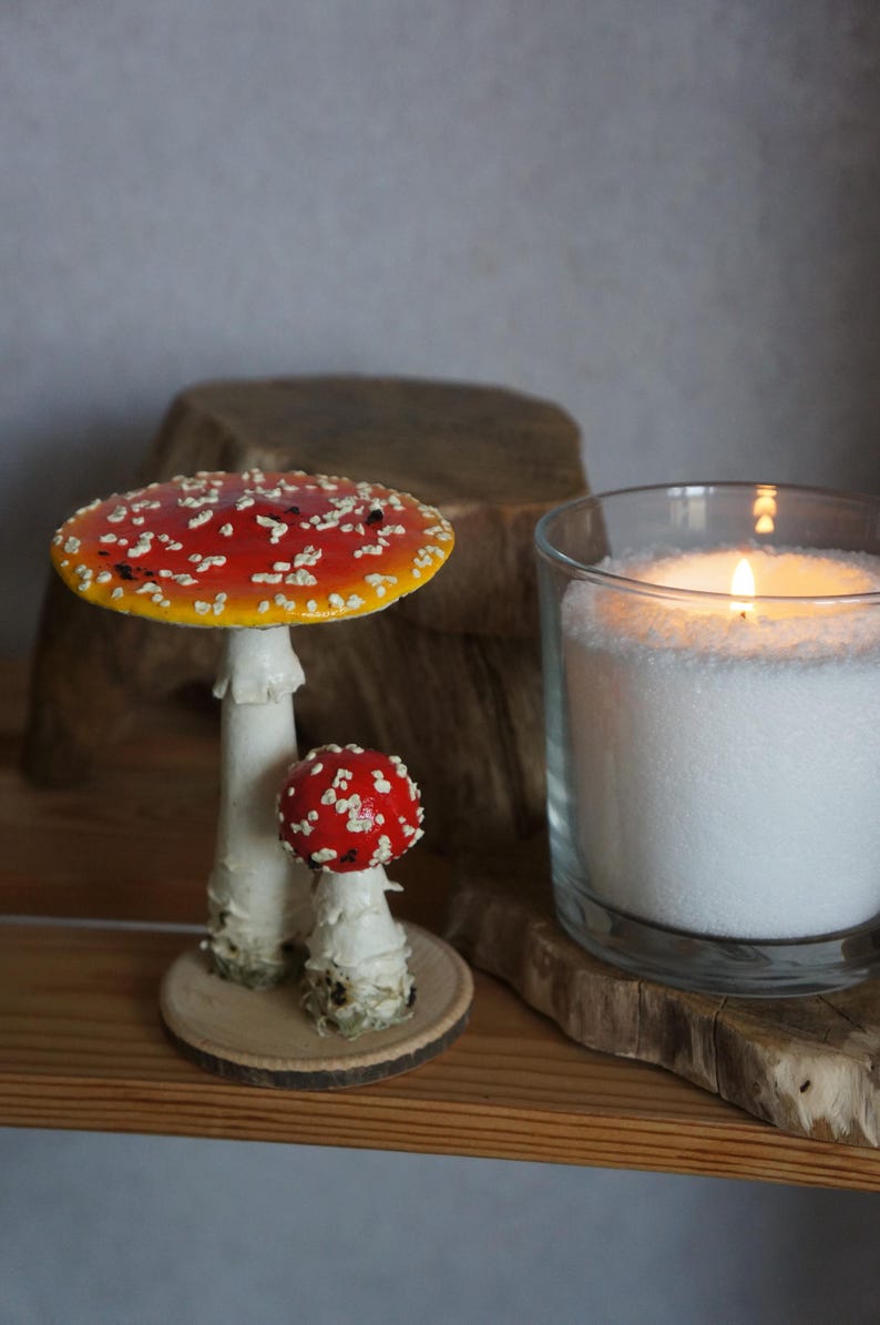 May include: Two decorative mushroom figurines with red caps and white spots, one large and one small, sit on a wooden surface. A lit candle in a glass jar is also present, creating a cozy, natural aesthetic.