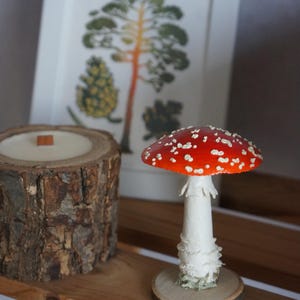 May include: A decorative arrangement featuring a red and white mushroom figurine, a wood-grain candle, and a framed botanical print. The mushroom has a white stem and a red cap with white spots. The candle is in a wood log container.