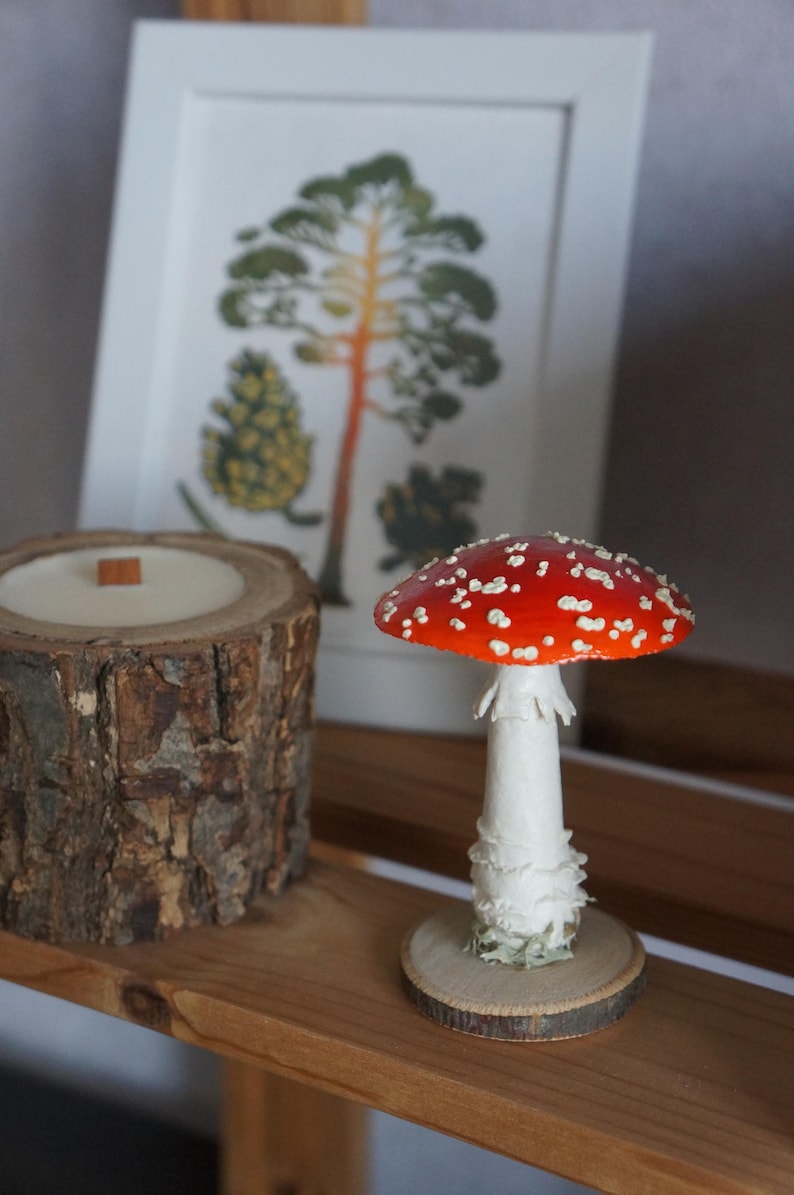 May include: A decorative arrangement featuring a red and white mushroom figurine, a wood-grain candle, and a framed botanical print. The mushroom has a white stem and a red cap with white spots. The candle is in a wood log container.