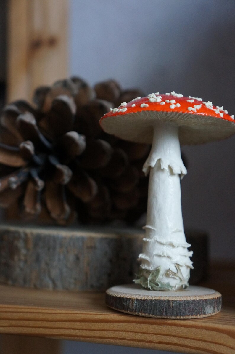 May include: A decorative mushroom figurine with a red cap dotted with white spots. The mushroom has a white stem and sits on a small wooden base. A pine cone is in the background.