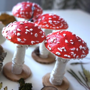 May include: A collection of handmade decorative mushrooms. The mushrooms have bright red caps with white spots, white stems, and are mounted on small wooden bases. The image shows several mushrooms of varying sizes.