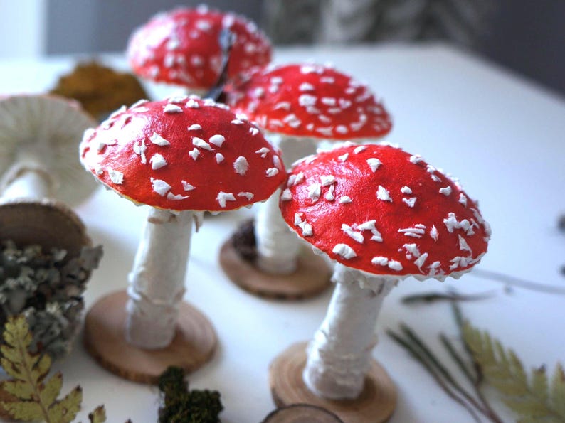 May include: A collection of handmade decorative mushrooms. The mushrooms have bright red caps with white spots, white stems, and are mounted on small wooden bases. The image shows several mushrooms of varying sizes.