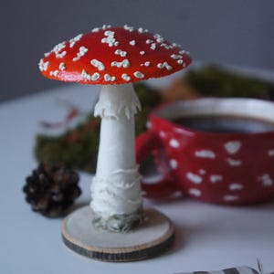 May include: A decorative mushroom figurine with a red cap and white spots, set on a wooden base. A red mug with white polka dots and a feather are in the background. The mushroom is approximately 4 inches tall.