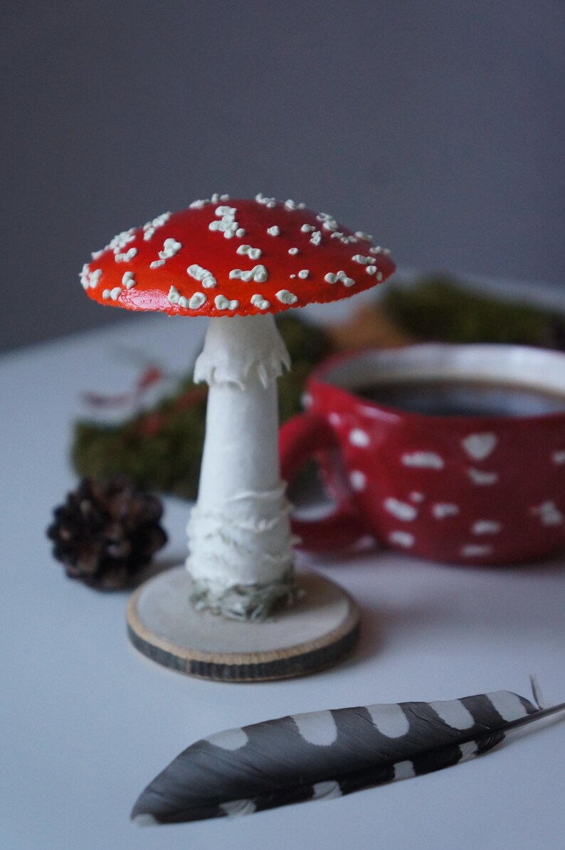 May include: A decorative mushroom figurine with a red cap and white spots, set on a wooden base. A red mug with white polka dots and a feather are in the background. The mushroom is approximately 4 inches tall.