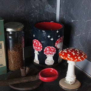 May include: A ceramic mug with a dark blue background and red interior, decorated with white stars and red and white mushroom illustrations. A small red bowl and a mushroom figurine are also present.