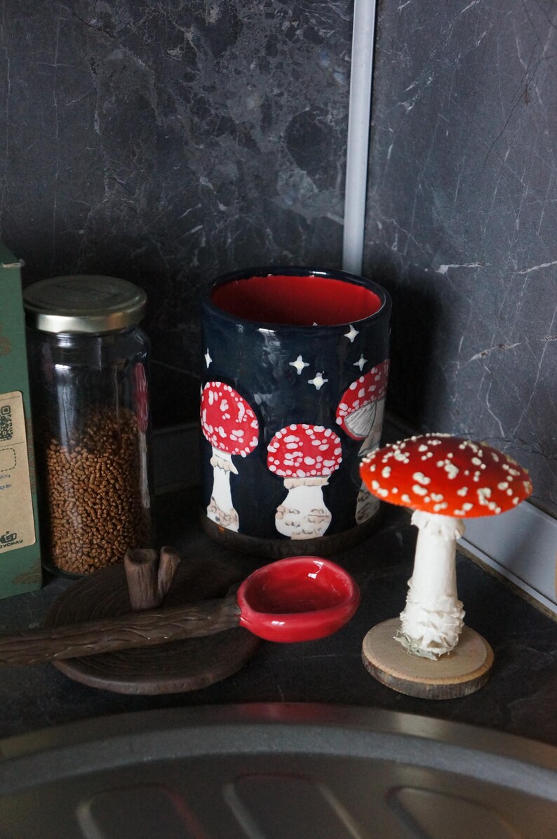 May include: A ceramic mug with a dark blue background and red interior, decorated with white stars and red and white mushroom illustrations. A small red bowl and a mushroom figurine are also present.