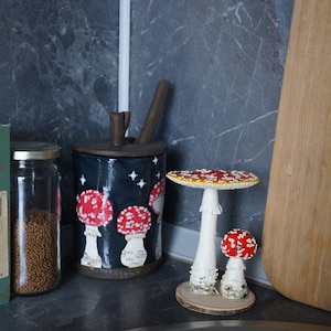 May include: A whimsical kitchen scene featuring a dark blue canister adorned with red and white mushroom illustrations. Two decorative mushroom figurines with red and white caps sit on a wooden base. A wooden cutting board and a jar are also visible.