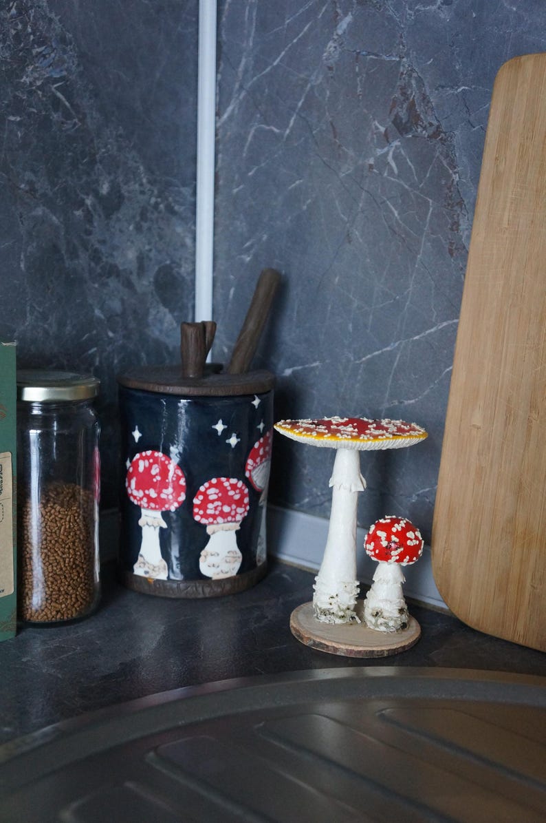 May include: A whimsical kitchen scene featuring a dark blue canister adorned with red and white mushroom illustrations. Two decorative mushroom figurines with red and white caps sit on a wooden base. A wooden cutting board and a jar are also visible.