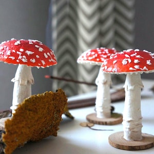 May include: Three decorative mushroom figurines with red caps and white spots. The mushrooms have white stems and are mounted on small wooden bases. A piece of textured, brown bark is in the foreground.