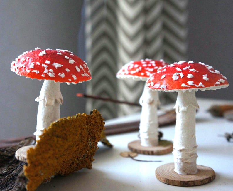 May include: Three decorative mushroom figurines with red caps and white spots. The mushrooms have white stems and are mounted on small wooden bases. A piece of textured, brown bark is in the foreground.
