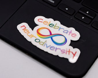 May include: A black laptop keyboard with a trackpad. A white sticker with the words "celebrate neurodiversity" in rainbow colors and an infinity symbol is affixed to the trackpad.