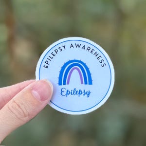 A circular sticker with a light purple background featuring a purple awareness rainbow in the middle with "Epilepsy" underneath. The words "Epilepsy Awareness" wrap around the top half of the sticker.