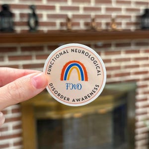 A circular sticker with a light orange background featuring a blue and orange awareness rainbow in the middle with "FND" underneath. The words "Functional Neurological Disorder Awareness" wrap around the design.