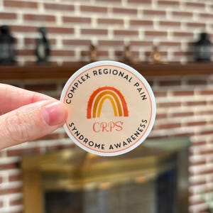 A circular sticker with a light orange background featuring an orange awareness rainbow in the middle with "CRPS" underneath. The words "Complex Regional Pain Syndrome Awareness" wrap around the design.