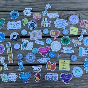 An assortment of colorful stickers and buttons featuring various designs and phrases related to chronic health conditions, disability, and neurodivergence. The stickers and buttons are arranged on a wood background. There are designs for conditions like Postural Orthostatic Tachycardia Syndrome (POTS), Gastroparesis, Ehlers-Danlos Syndrome (EDS), Mast Cell Activation Syndrome (MCAS), food allergies, eating disorder recovery, mental health, and more.