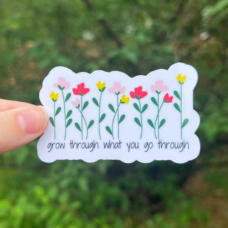 A white sticker with a row of yellow, pink, and red flowers with green stems. The text below the flowers reads "grow through what you go through" in black text