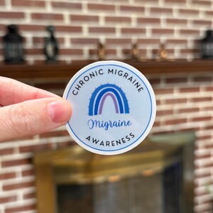 A circular sticker with a light purple background featuring a purple awareness rainbow in the middle with "Migraine" underneath. The words "Chronic Migraine Awareness" wrap around the design.