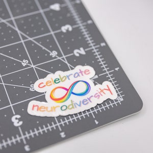 A rainbow sticker that says "celebrate neurodiversity" featuring a rainbow infinity sign on a gray cutting mat. The mat shows the sticker is about 2.5 inches across and 1.5 inches tall