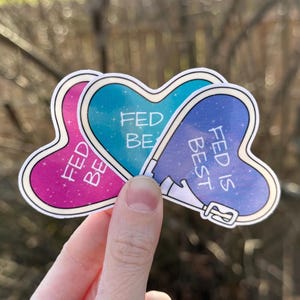 May include: Three heart-shaped stickers with the words "FED BE" and "FED IS BEST" in white. The stickers have a white border and are in shades of pink, teal, and blue with a starry background.