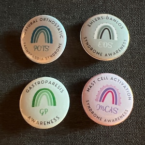 May include: Four round buttons with rainbow designs and text. The buttons are white with a light blue, gray, green, and pink background. The text on the buttons reads "Postural Orthostatic Tachycardia Syndrome", "Ehlers-Danlos Syndrome", "Gastroparesis", and "Mast Cell Activation Syndrome".