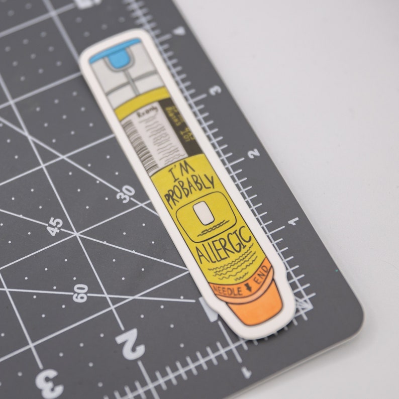 A sticker of a yellow and orange epinephrine Epi-Pen auto-injector. The sticker says "I'm Probably Allergic" near the orange needle end of the device. The sticker is shown on a gray cutting mat showing it is roughly one inch across and four inches tall.