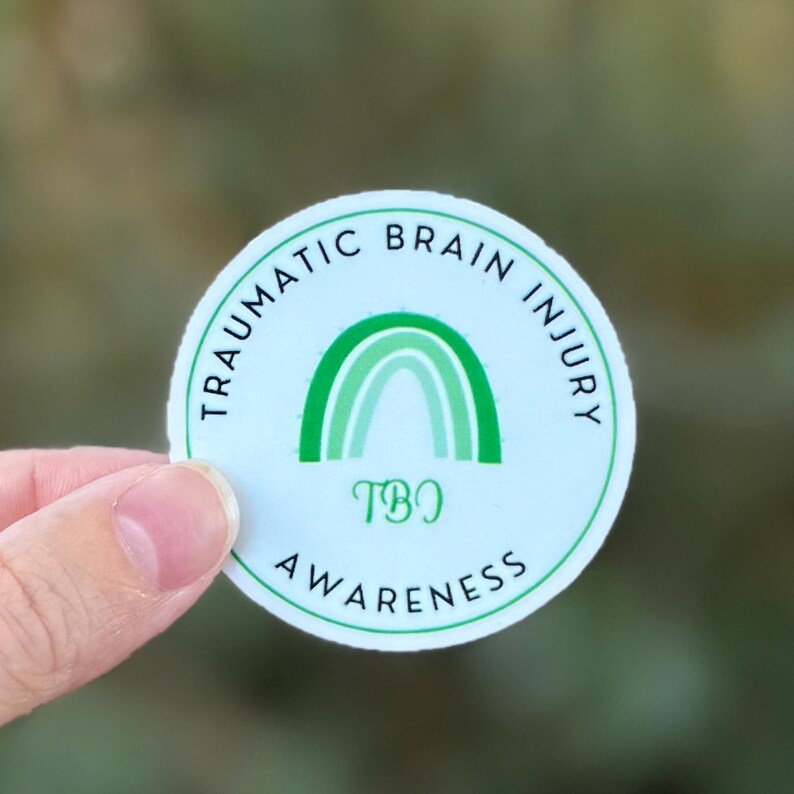 A circular sticker with a light green background featuring a green awareness rainbow in the middle with "TBI" underneath. The words "Traumatic Brain Injury Awareness" wrap around the design.