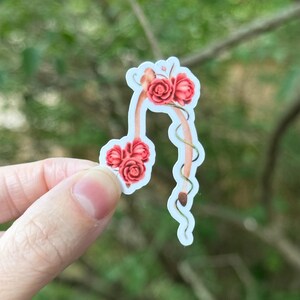 May include: A sticker featuring a floral design with red roses and green vines. The sticker has a light blue border and a tan stem-like shape. The roses are clustered in three groups, with the largest group at the top.
