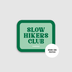 May include: A square iron-on patch with rounded corners. The patch features the text "SLOW HIKERS CLUB" in dark green, with "CANADA" below. A white circle with "IRON-ON PATCH" is on the right.