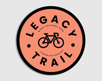 May include: A black and orange round sticker with the text "LEGACY TRAIL" in a circle around a bicycle silhouette. The text "BANFF NATIONAL PARK" and "CANADIAN ROCKIES" are also included in the design.