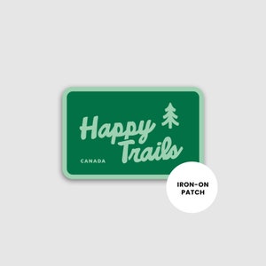 May include: A green rectangular iron-on patch with the words "Happy Trails" in a whimsical font, a tree graphic, and the word "CANADA". A white circle with the words "IRON-ON PATCH" is below the patch.