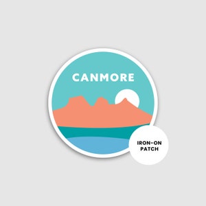 May include: A round, blue and white iron-on patch with the text "Canmore" and a silhouette of mountains and a sun. The text "Iron-on Patch" is also included.