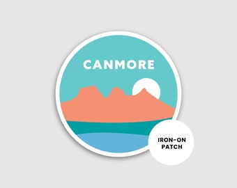 May include: A round, blue and white iron-on patch with the text "Canmore" and a silhouette of mountains and a sun. The text "Iron-on Patch" is also included.