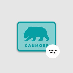 May include: An iron-on patch featuring a teal bear silhouette on a light blue rectangular background. The word "CANMORE" is printed in teal below the bear. A white circle with "IRON-ON PATCH" is on the right.