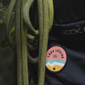 May include: A green and black rope is wrapped around a black backpack with a pink and yellow patch that says "Lake Louise 4200 Acres".